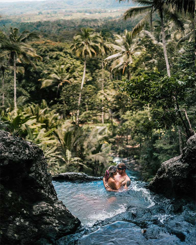 A couple swim in a waterfall with palm trees and jungle in the background.
