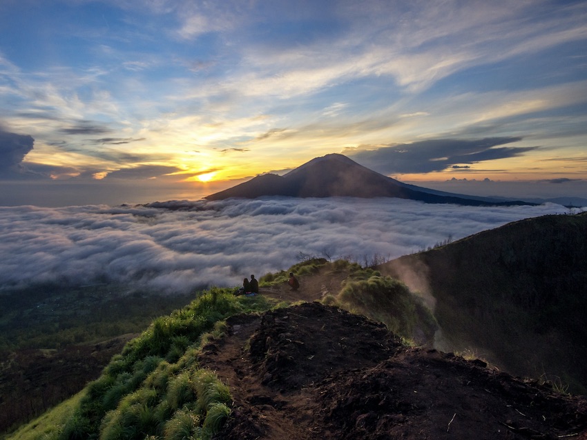 The sun rises over Mt. Agung as 2 people watch from the top of Mt. Batur.