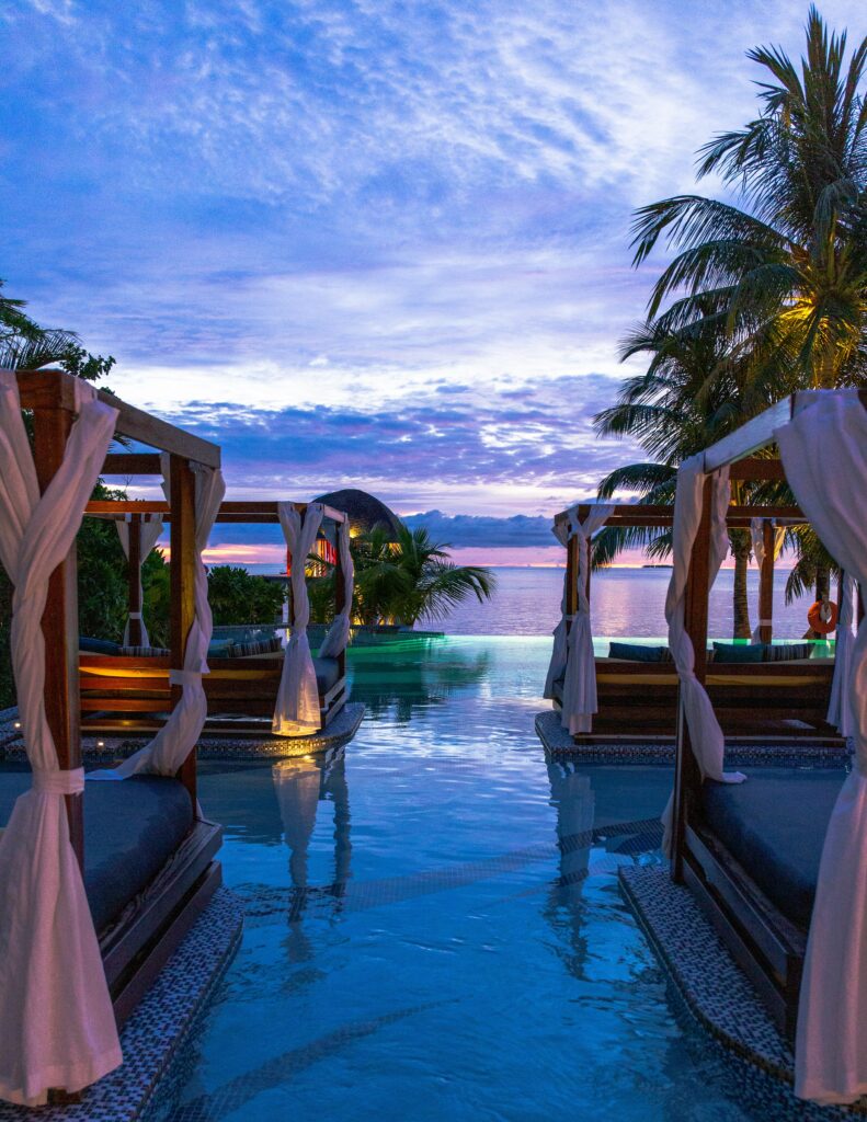 Day beds with canopy are in an infinity pool in Bali with a pink sunset in the background.