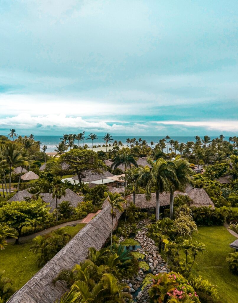 A resort in Fiji has tall palm trees, thatched roof huts and views of white sand beaches under the white clouds.