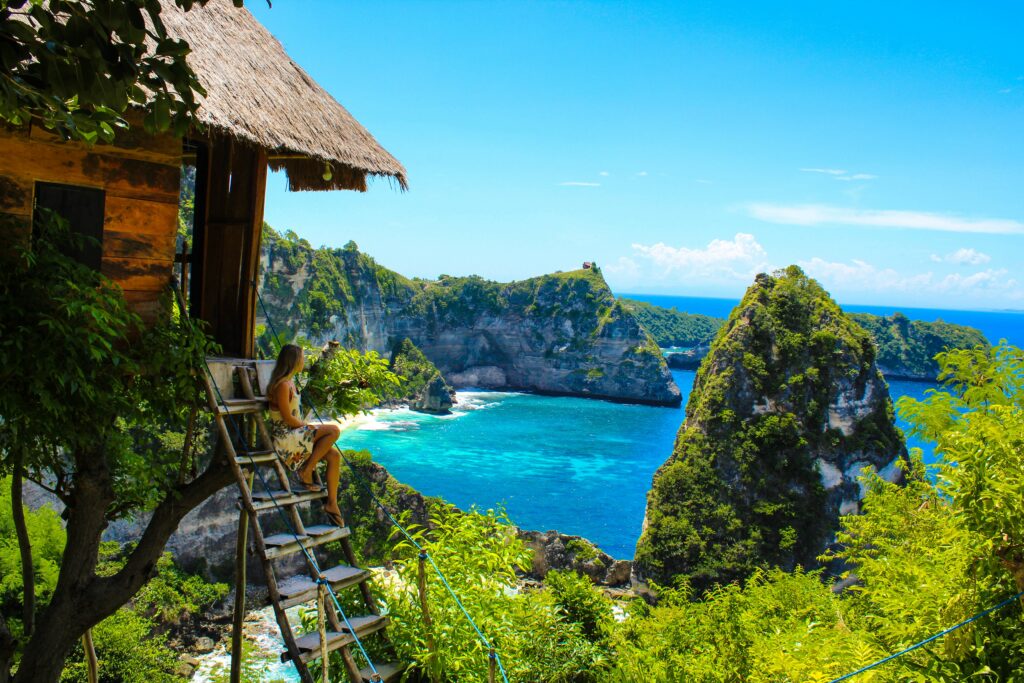 A woman sits on the steps of a treehouse looking at rock formations and the blue ocean.